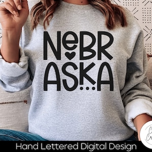 May include: A gray sweatshirt with the text "NEBR ASKA..." in black lettering with a small heart between the words "NE" and "BR".