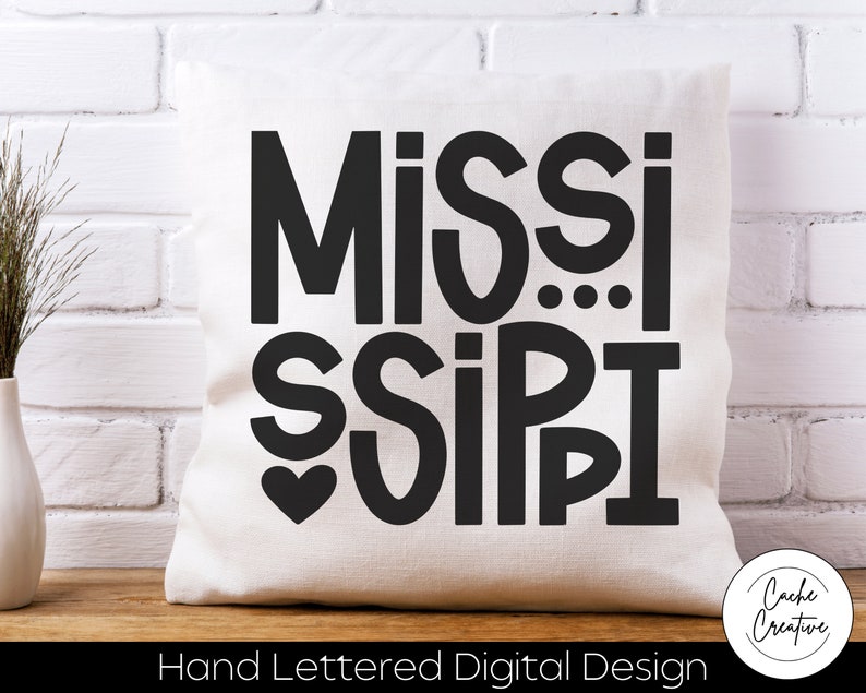 May include: White pillow with black text that reads "MISSISSIPPI" with a heart in the middle. The text is in a bold, hand-lettered style. The pillow is on a white brick background. The text "Hand Lettered Digital Design" is below the pillow.