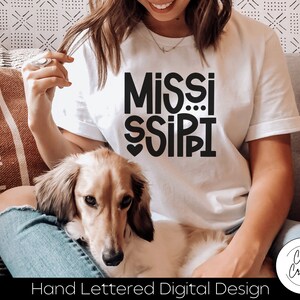 May include: A white t-shirt with a black hand-lettered design that says "MISSISSIPPI" with a heart in the middle of the "I". The shirt is being worn by a person sitting on a couch with a dog.
