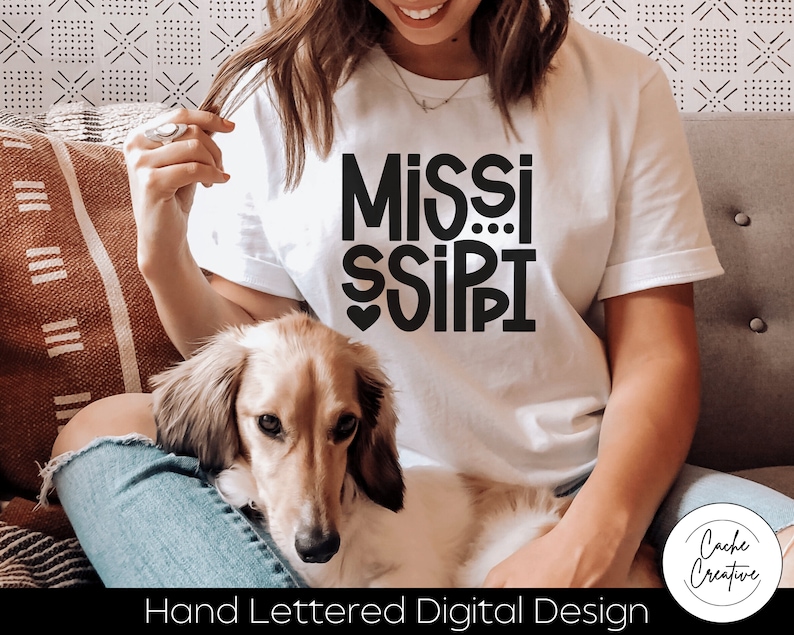 May include: A white t-shirt with a black hand-lettered design that says "MISSISSIPPI" with a heart in the middle of the "I". The shirt is being worn by a person sitting on a couch with a dog.