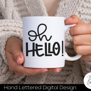 Puede incluir: Una taza de cerámica blanca con el texto negro escrito a mano "oh HELLO!" y un pequeño corazón integrado en la letra O. La taza tiene un asa estándar en el lado derecho y un cuerpo cilíndrico liso con un diseño limpio y sencillo.
