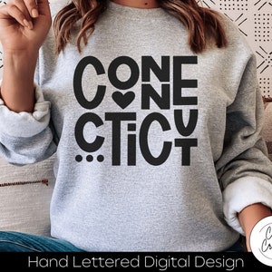 Peut inclure: Sweat-shirt gris avec un texte noir qui dit "CONNE CTICUT" avec un petit cœur entre les mots "CON" et "NE".