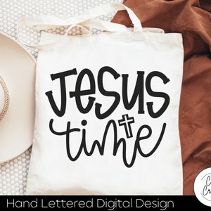 Könnte beinhalten: Weiße Einkaufstasche mit schwarzer Aufschrift "Jesus time" mit einem kleinen Kreuz über dem Wort "time". Die Tasche liegt auf einer weißen und braunen strukturierten Oberfläche.
