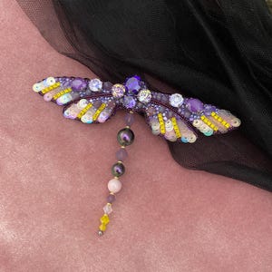May include: A purple and yellow dragonfly brooch with iridescent sequins and beads. The dragonfly has a long tail of purple, pink, and black beads.