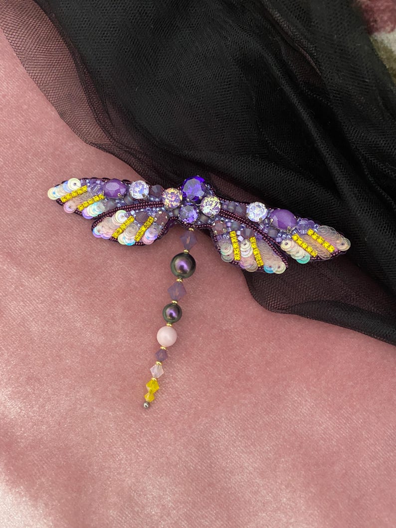 May include: A purple and yellow dragonfly brooch with iridescent sequins and beads. The dragonfly has a long tail of purple, pink, and black beads.