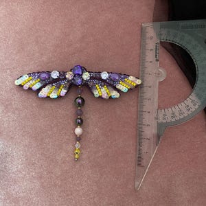 May include: A purple and yellow beaded dragonfly brooch with a dangling chain of beads. The brooch is approximately 6 cm long.