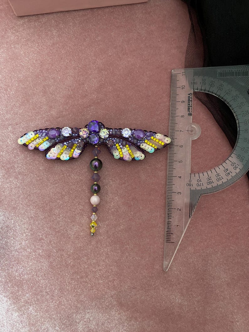 May include: A purple and yellow beaded dragonfly brooch with a dangling chain of beads. The brooch is approximately 6 cm long.