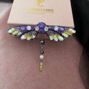 May include: A purple and yellow dragonfly brooch with iridescent sequins and beads. The dragonfly has a large purple gemstone in the center of its body and a dangling pearl and crystal chain.