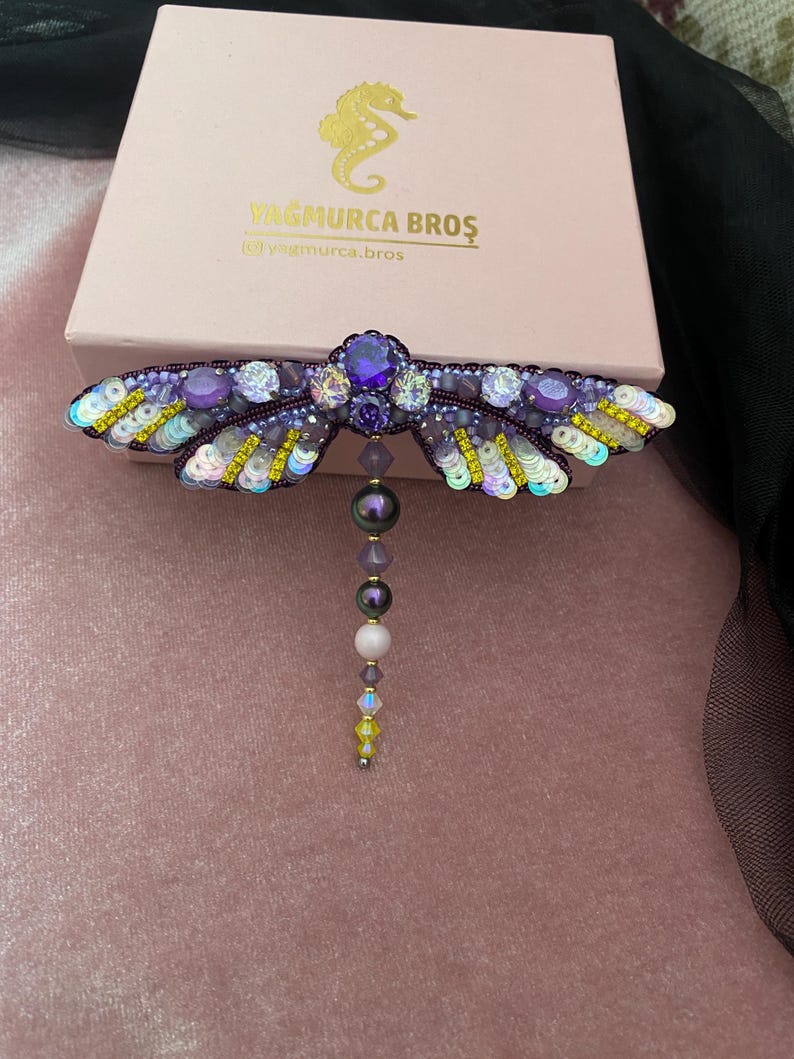 May include: A purple and yellow dragonfly brooch with iridescent sequins and beads. The dragonfly has a large purple gemstone in the center of its body and a dangling pearl and crystal chain.