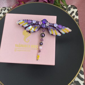 May include: A purple and gold dragonfly brooch with a beaded tail. The brooch is on a pink box with the text "YAGMURCA BROS" and "@yagmurca_bros".