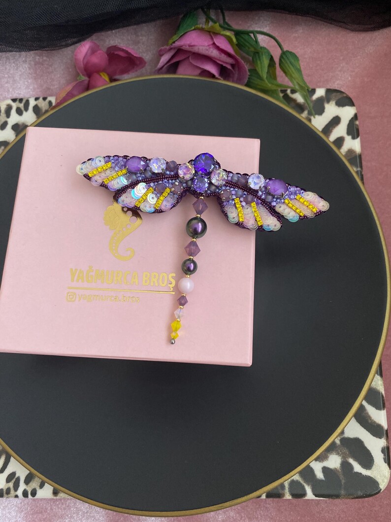 May include: A purple and gold dragonfly brooch with a beaded tail. The brooch is on a pink box with the text "YAGMURCA BROS" and "@yagmurca_bros".