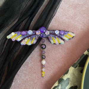 May include: A purple and yellow beaded dragonfly brooch with a dangling beaded tail. The dragonfly is made with iridescent beads and sequins.