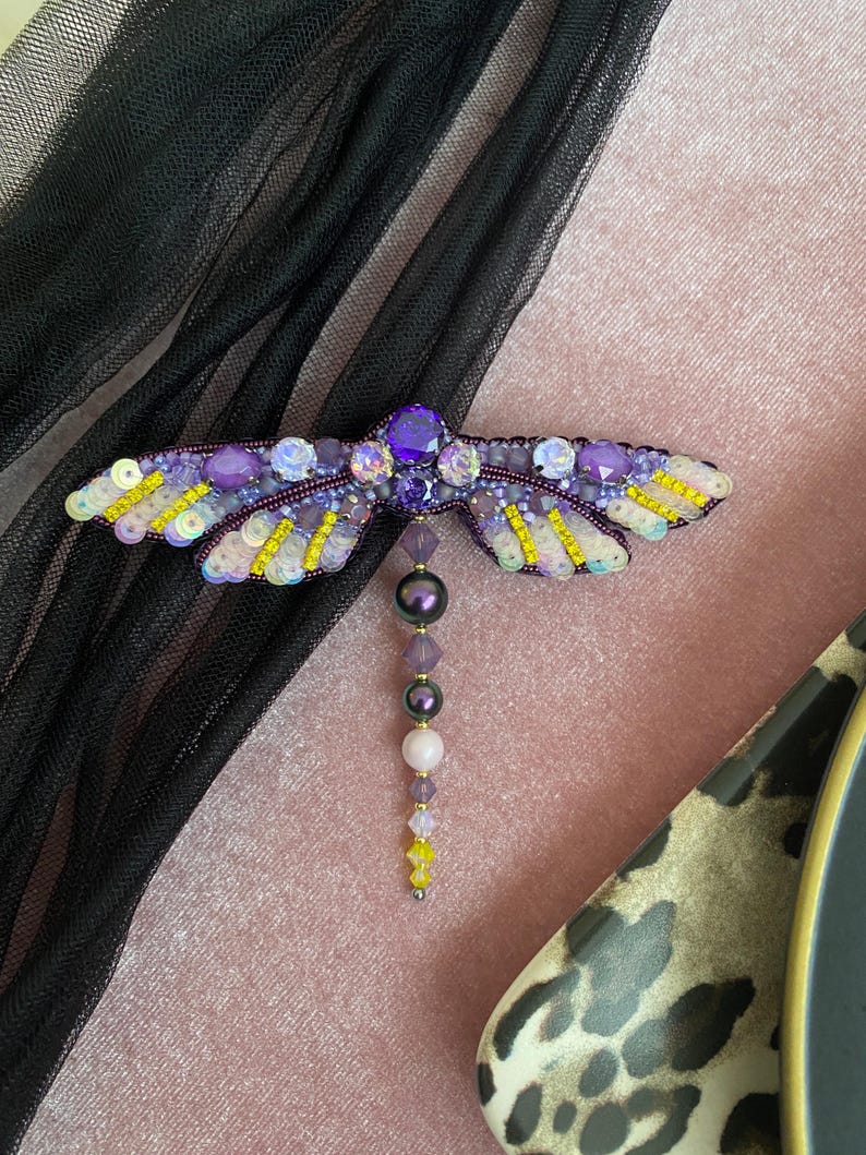 May include: A purple and yellow beaded dragonfly brooch with a dangling beaded tail. The dragonfly is made with iridescent beads and sequins.