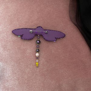 May include: Purple fabric dragonfly brooch with a beaded dangle. The beads are purple, white, and yellow.