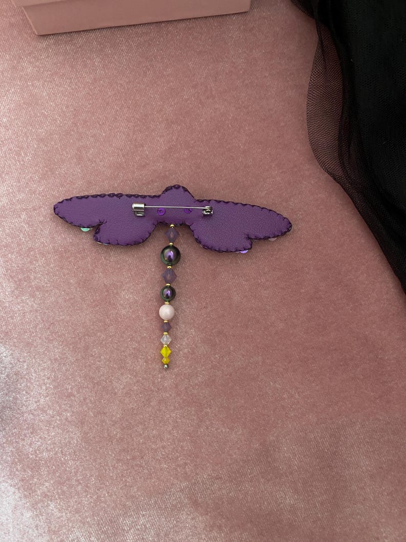 May include: Purple fabric dragonfly brooch with a beaded dangle. The beads are purple, white, and yellow.