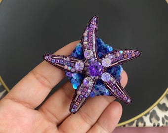 Puede incluir: Broche de estrella de mar artesanal con un diseño de cuentas moradas y rosas. El broche presenta una gema central morada rodeada de gemas y cuentas más pequeñas, sobre una base de lentejuelas azules. El broche se sostiene en una mano.