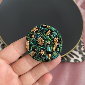May include: A round green and gold beaded brooch with a mosaic pattern.