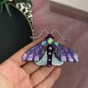 May include: A purple and silver moth brooch with iridescent sequins and beads. The moth has a velvet body and wire antennae.