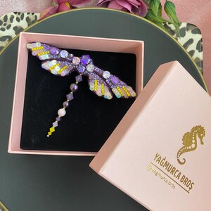 May include: A purple and yellow dragonfly brooch with beaded wings and a dangling chain of beads. The brooch is in a pink box with the text "Yağmurca Bros" and a social media handle.