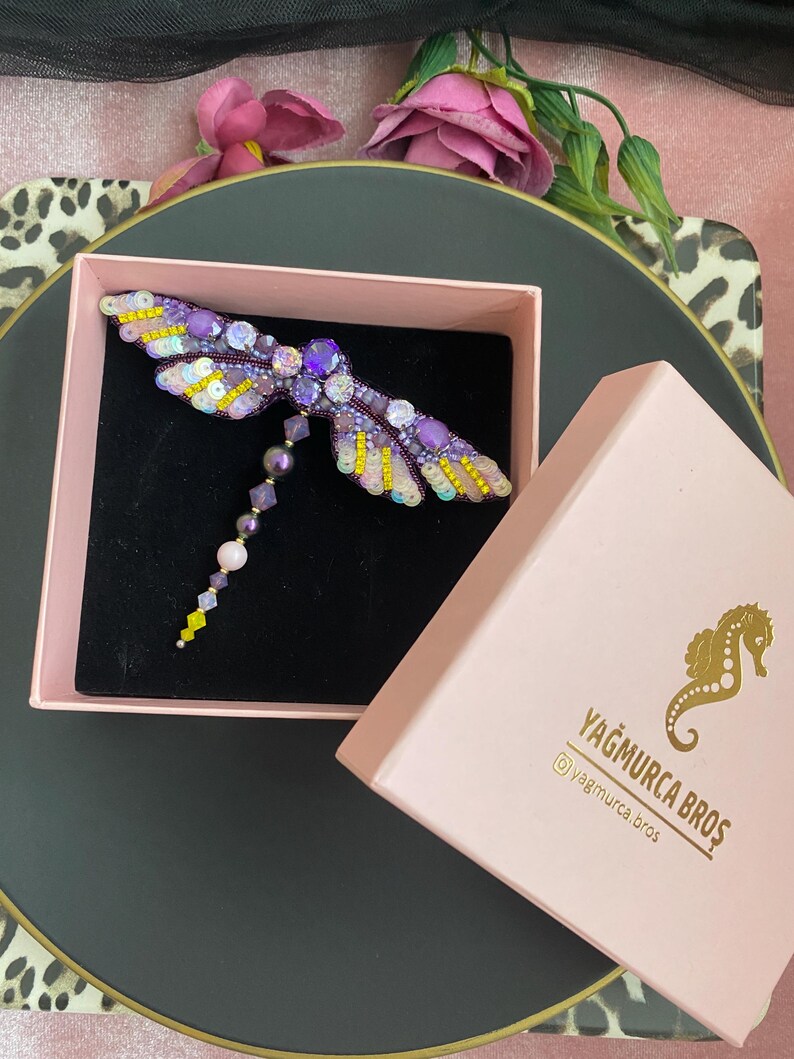 May include: A purple and yellow dragonfly brooch with beaded wings and a dangling chain of beads. The brooch is in a pink box with the text "Yağmurca Bros" and a social media handle.