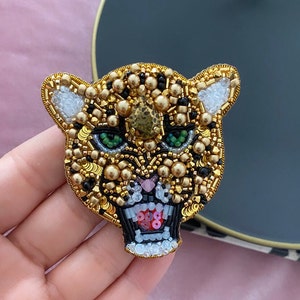 Puede incluir: Un broche de jaguar de cuentas doradas, negras y verdes con una expresión feroz. El broche está hecho con cuentas pequeñas y lentejuelas, creando un diseño detallado y texturizado.