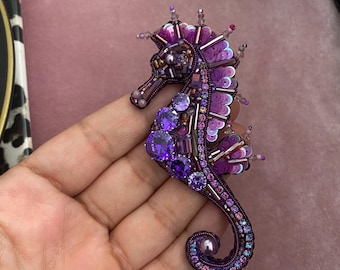 Puede incluir: Broche de caballito de mar hecho a mano en tonos morados, adornado con cuentas, lentejuelas y cristales. El broche presenta un diseño detallado con una cola rizada y una superficie texturizada, creando un efecto brillante. El caballito de mar mide aproximadamente 7.6 cm de largo.