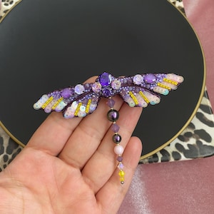 May include: A purple and yellow dragonfly brooch with iridescent sequins and beads. The dragonfly has a long, beaded tail.