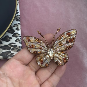 May include: A detailed butterfly brooch, featuring a complex bead design in brown, gold, and cream hues. The wings are adorned with beads and crystals, with a large, clear crystal at the centre. The brooch has a bronze-coloured metal frame.
