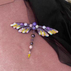 May include: A purple and yellow beaded dragonfly brooch with a beaded tail. The dragonfly is on a pink velvet background.
