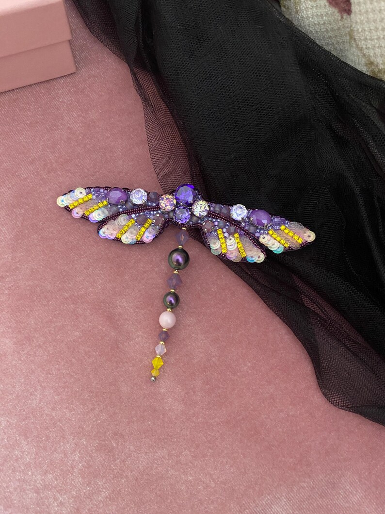 May include: A purple and yellow beaded dragonfly brooch with a beaded tail. The dragonfly is on a pink velvet background.