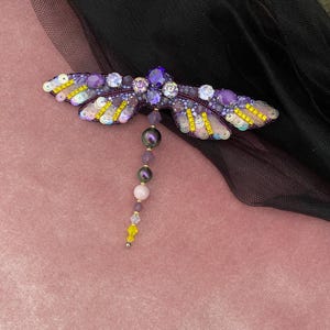 May include: A purple and yellow dragonfly brooch with iridescent sequins and beads. The dragonfly has a long tail of purple, pink, and yellow beads.