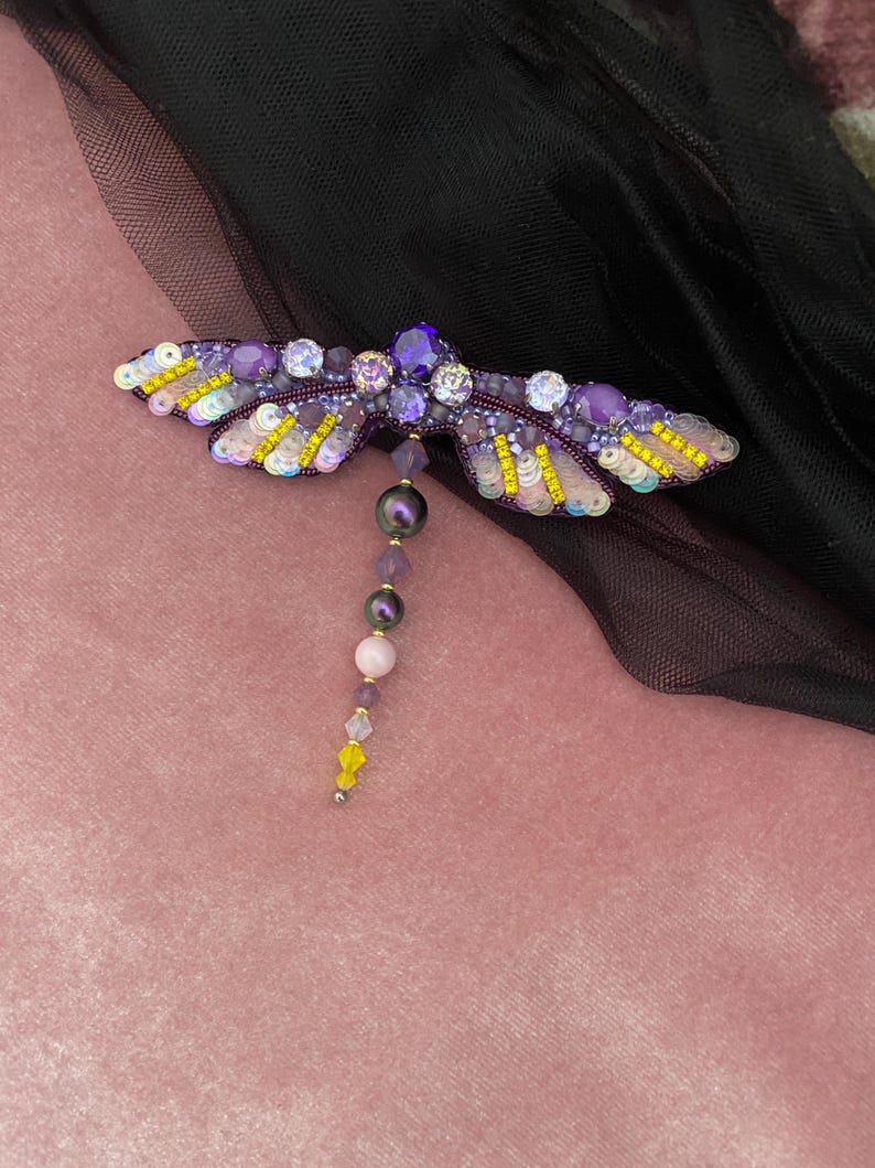 May include: A purple and yellow dragonfly brooch with iridescent sequins and beads. The dragonfly has a long tail of purple, pink, and yellow beads.