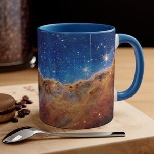 May include: A blue ceramic coffee mug with a colorful nebula image printed on the front. The mug has a white interior and a blue handle.