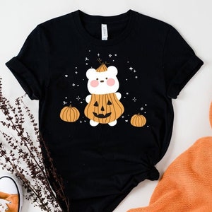 Op de afbeelding: Zwart T-shirt met een witte beer die een pompoenkostuum draagt. De beer heeft een schattig gezicht en is omringd door kleine pompoenen. Het shirt heeft een Halloween-thema.