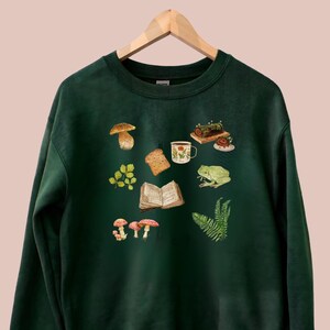 Puede incluir: Una sudadera verde oscuro con una escena de bosque caprichosa impresa en la parte delantera. El diseño presenta hongos, una rana, un libro, una taza de té, una rebanada de pan y un helecho.