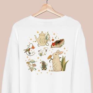 Puede incluir: Sudadera blanca con un dise&ntilde;o caprichoso que presenta una tetera, un libro, un conejo, champi&ntilde;ones, un cristal y un colibr&iacute;. El dise&ntilde;o est&aacute; rodeado de estrellas doradas.