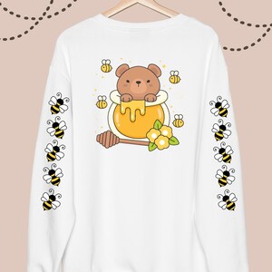 Puede incluir: Sudadera blanca con un oso de dibujos animados en un tarro de miel en la espalda. Las mangas tienen un patr&oacute;n repetitivo de abejas de dibujos animados.