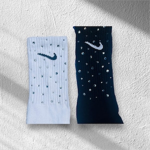 May include: A pair of athletic socks, one white and one navy blue, each adorned with sparkling embellishments. Both socks feature the Nike swoosh logo. The white sock has a dark teal logo, while the navy sock has a white logo.