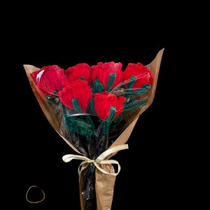 May include: A bouquet of red roses wrapped in brown paper and clear cellophane, tied with a gold ribbon. The roses have green stems and leaves. The background is solid black.