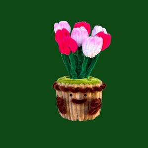 Pode incluir: Um artesanato artesanal e caprichoso com um vaso de flores com um rosto sorridente, segurando um buquê de tulipas rosa e vermelhas feitas com limpadores de cachimbo. O vaso é bege com detalhes marrons e 'grama' verde.