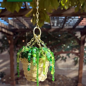 May include: A hanging basket with a woven design, filled with green foliage and beaded accents. The basket is suspended by a gold chain. The text "Plant 2" is in the upper left corner.