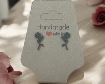 Puede incluir: Pendientes de metal gris con forma de figuras de palo corriendo, presentados en una tarjeta gris. La tarjeta tiene un agujero troquelado y el texto "Handmade with love".