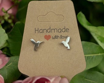 Könnte beinhalten: Ohrstecker mit Kolibri-Motiv aus Silber auf einer braunen Karte mit dem Text "Handmade with love". Der Hintergrund zeigt rosa Blüten und grüne Blätter.