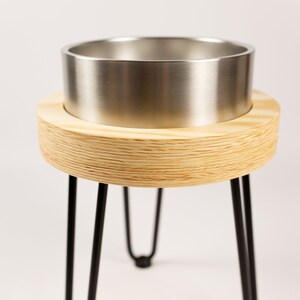 May include: A wooden plant stand with a metal planter. The stand has three black metal legs and a round wooden top. The planter is made of stainless steel and has a smooth, shiny finish.