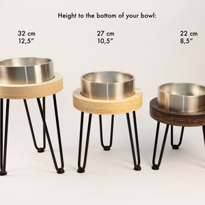 May include: Three elevated pet bowls with different heights: 32 cm, 27 cm, and 22 cm. The bowls are made of stainless steel and are set on wooden stands with black metal legs.