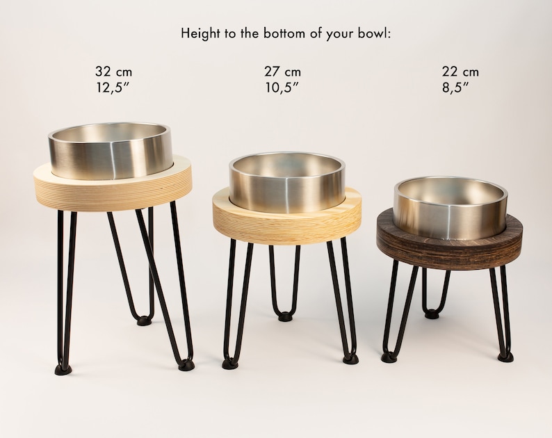 May include: Three elevated pet bowls with different heights: 32 cm, 27 cm, and 22 cm. The bowls are made of stainless steel and are set on wooden stands with black metal legs.