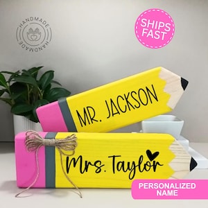 May include: Two personalized wooden pencil-shaped signs. One is yellow with "MR. JACKSON" in black, the other is yellow with "Mrs. Taylor" and a heart. Both have pink erasers and gray bands. A bow adorns the second pencil. The signs are handmade.