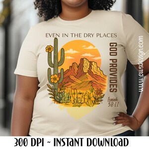 May include: Cream-colored t-shirt featuring a desert landscape design with a cactus, mountains, and a setting sun. The text reads "Even in the dry places, God provides" with a Bible verse reference. The design is in shades of orange, yellow, and green.