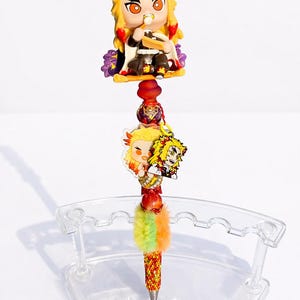 May include: A decorative pen featuring a character figurine with yellow hair and red accents. The pen has a beaded design with a colorful pom-pom and is displayed in a clear pen holder.