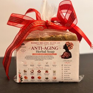 Può includere: Una saponetta rettangolare alle erbe anti-età di Women Breathe Again Co. avvolta in plastica trasparente con un fiocco rosso. L'etichetta presenta testo, un'illustrazione a silhouette e icone che descrivono ingredienti come olio di cocco e burro di karité.